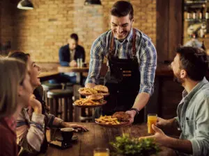 Happy waiter serving food to group of cheerful friends in a pub