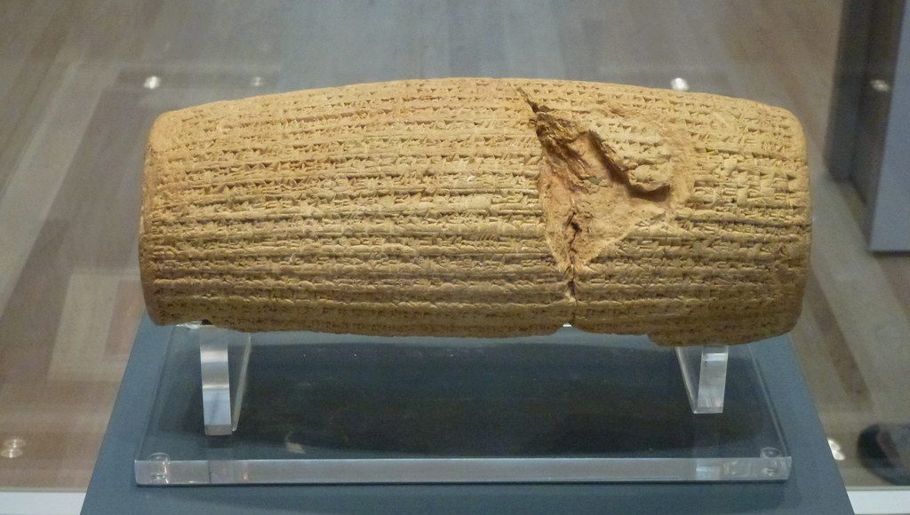 Image of the Cyrus Cylinder at the British Museum, featuring ancient Persian cuneiform inscriptions on a clay barrel